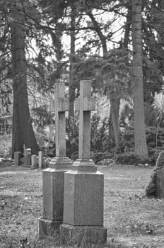Two old gravestones with crosses stand in a peaceful, wooded cemetery. The weathered stones and the natural surroundings create a melancholy atmosphere.