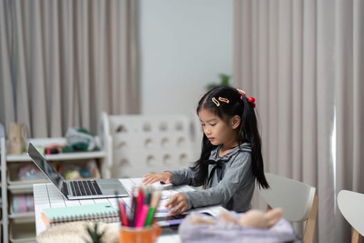 A young girl is focused on her studies at home, working diligently on her laptop in a cozy study space.