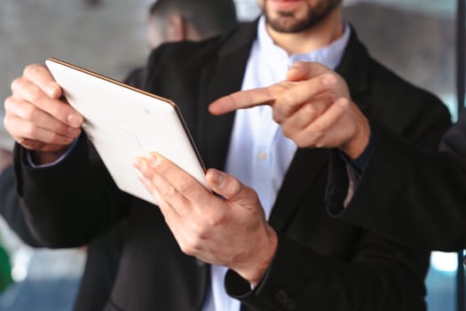 Unrecognizable businessmen in formal suits using a tablet during a meeting. One man points to the screen in a modern workspace, emphasizing teamwork, technology, and collaboration.