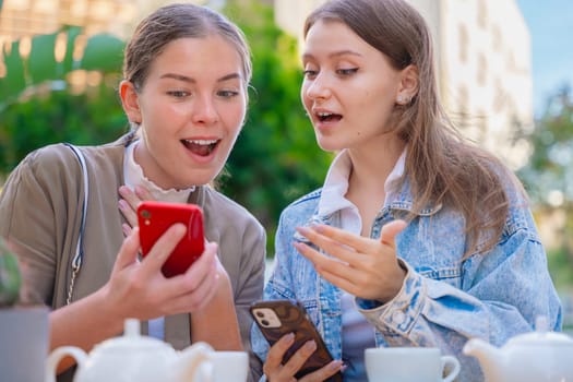 Two happy friends women sitting at an outdoor cafe, using smartphones and reacting with curiosity and surprise. Surrounded by teapots and cups, they share a modern and relaxed social moment together.