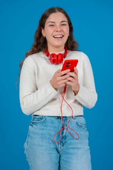Happy young caucasian woman wearing red headphones around her neck,smiling while looking at her smartphone looking at camera.The blue background highlights her joyful and engaged expression.Vertical