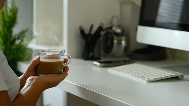 Close up of person holding a steaming cup of coffee while working in a home workspace.