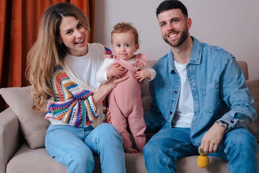 Portrait Happy young parents with their baby sitting on a sofa and smiling at the camera. Cozy family moment indoors with love, warmth, and togetherness in a relaxed home environment.