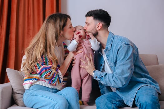 Happy parents lovingly kiss their baby on both cheeks while sitting on a cozy sofa at home. The scene captures warmth, tenderness, and family bonding in a casual indoor environment.