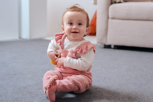 Cute baby in pink overalls sitting on a soft carpet, smiling and holding a small toy while enjoying playtime indoors in a cozy home environment with soft lighting and neutral background.