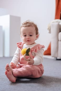 Cute baby in pink overalls sitting on a soft carpet, smiling and holding a small toy while enjoying playtime indoors in a cozy home environment with soft lighting and neutral background.