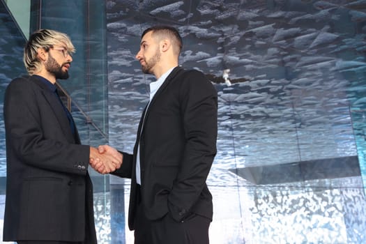 Two professional businessmen in suits shaking hands inside a modern glass office. Business deal, agreement, or partnership in a corporate indoor setting with contemporary design. Copy space