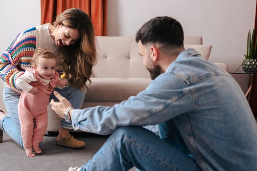 Mother and father encourage their baby girl as she takes her first steps on the carpet. A heartwarming family moment filled with love, support, and early childhood development at home.