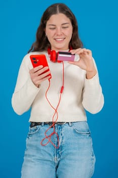 Happy young woman in a white sweater and red headphones smiling while showing a credit card to the camera and holding a red smartphone, against a blue background, symbolizing online shopping. Vertical