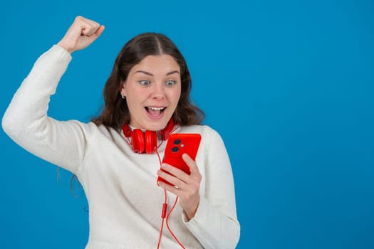 Happy young woman wearing red headphones around her neck, clenching her fist in excitement while looking at her smartphone. Her joyful reaction stands out against the blue background.Copy space