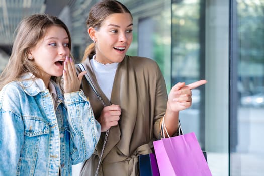 Two happy Caucasian women holding shopping bags and pointing at a store window display. Candid and joyful urban moment during a day of shopping and friendship in the city.