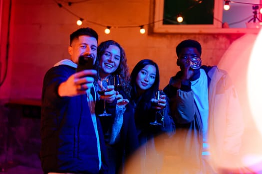 Four friends gather in a lively outdoor space, holding glasses and enjoying each other's company under colorful string lights at night.