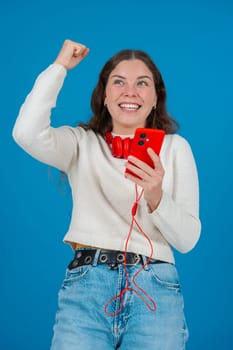 Happy young woman wearing red headphones around her neck, clenching her fist in excitement while looking at her smartphone. Her joyful reaction stands out against the blue background.Vertical