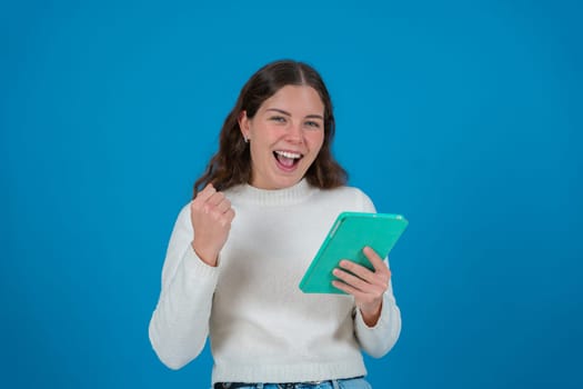 Happy young woman in a white sweater raising her fist in celebration while holding a green tablet and smiling at the camera, against a blue background, symbolizing success, joy, and digital life.