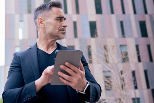 Businessman using a tablet outdoors in a modern city while looking away. He wears a black suit and smartwatch. Concept of technology, focus, mobility, and digital professional lifestyle.