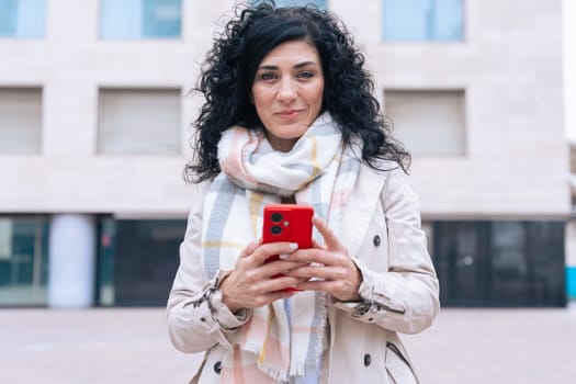 Happy caucasian woman standing outdoors in urban setting,wearing coat and scarf,using her red smartphone.She is smiling and relaxed,enjoying a casual moment on a cold day in the city looking at camera