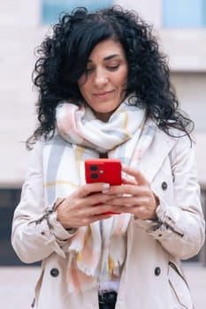 Serious caucasian woman standing outdoors in urban setting, wearing coat and scarf, using her red smartphone. She is serious and relaxed, enjoying a casual moment on a cold day in the city. Vertical