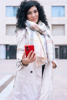 Happy caucasian woman standing outdoors in urban setting,wearing coat and scarf,using her red smartphone.She is smiling and relaxed,enjoying a casual moment on a cold day in the city looking at camera