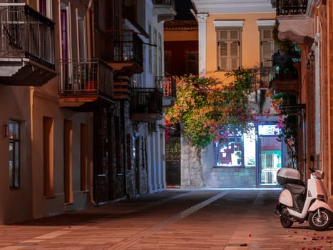 Beautiful night view of alleyway in Nafplio, Greece, at night. Bougainvillaea flowers and vines, scooter parked by doorway. Lights of some cafe at the end of the street.