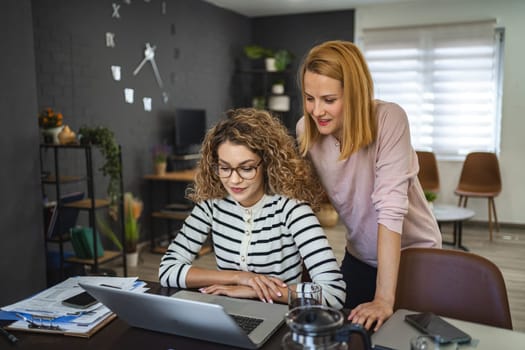 Two female friends are talking together in a video call on a laptop at the table in the living room.