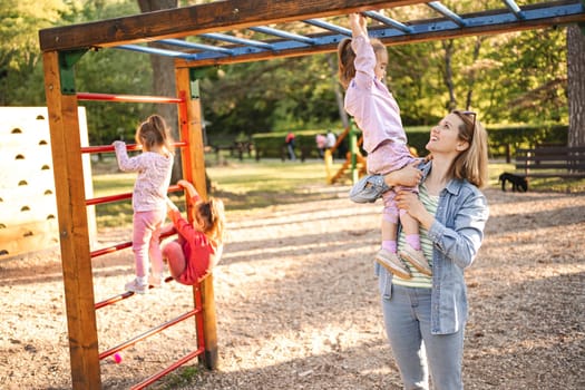 Smiling mom helps her cute daughter climb the playground.