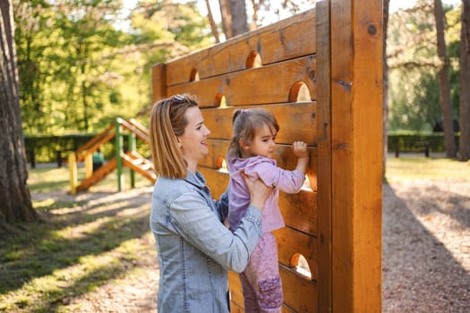 Smiling mom helps her cute daughter climb the playground.