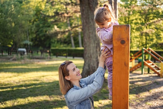 Smiling mom helps her cute daughter climb the playground.