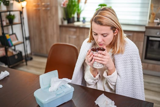 A woman enjoys a comforting drink while wrapped in a blanket at home.