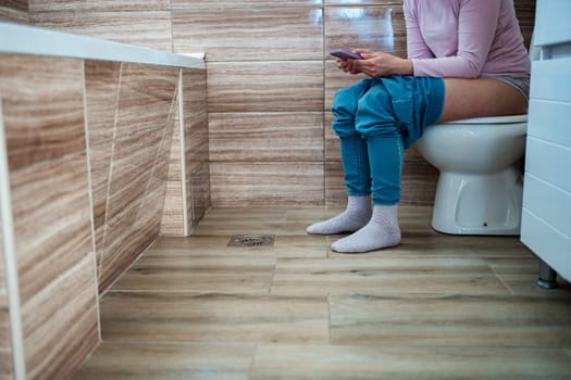 Individual relaxes on toilet, engaged with smartphone in cozy bathroom.