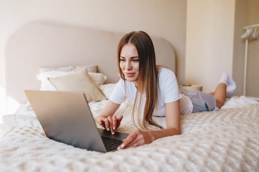 A young woman lies on her stomach on a bed, focused on her laptop. Sunlight streams into the cozy bedroom, creating a warm ambiance as she types away.