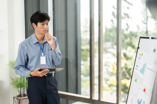 A young professional reflects on business data displayed on a whiteboard, holding a tablet in a modern office.