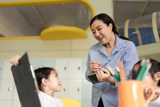 A teacher engages with a young student, discussing learning materials and encouraging participation in a bright classroom setting.