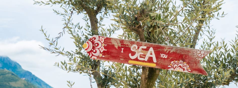 Wooden road sign on green olive tree with saying I SEA YOU joke pun. Concept of summer holiday vacation on seacoast. Idea of tropical travel tourism taking a break wellbeing slowing life.