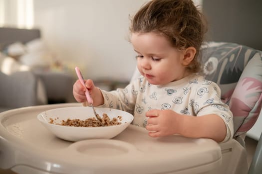 Cute little girl with curly hair sitting in a baby chair, enjoying oatmeal in a bright kitchen. Childhood mealtime moments.