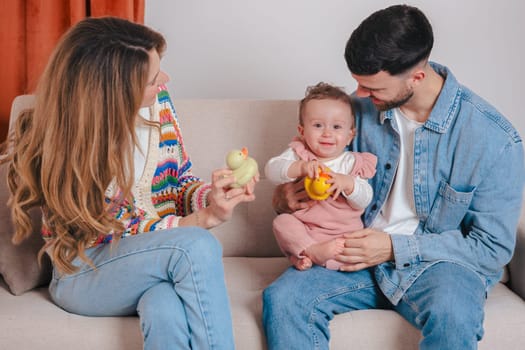 Smiling parents enjoy playtime with their baby girl who holds toys while sitting on a sofa. A joyful family bonding moment filled with love, color, and warmth in a cozy home setting.