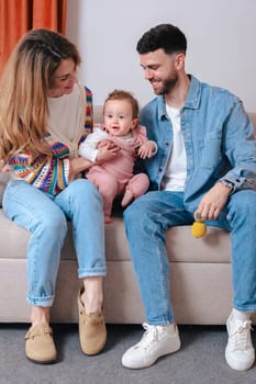 Vertical portrait Smiling parents enjoy playtime with their baby girl who holds toys while sitting on a sofa. A joyful family bonding moment filled with love, color, and warmth in a cozy home setting.