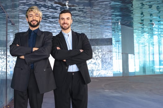 Two confident male professionals in suits standing with crossed arms, smiling at the camera inside a modern office. Corporate success, teamwork, and leadership in business environment. Copy space