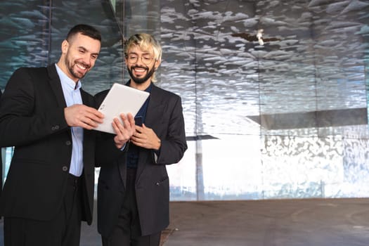 Two young businessmen in formal suits ,looking at a tablet and smiling while working together in a stylish modern office with reflective surfaces and a professional atmosphere. Copy space