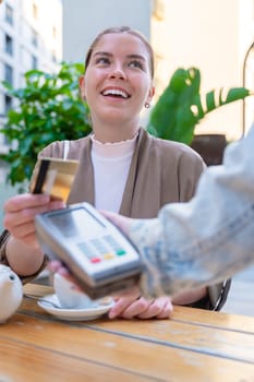 Caucasian woman smiling as she pays with a credit card using contactless technology at an outdoor cafe. A casual urban moment reflecting convenience, lifestyle, and modern financial habits. Vertical