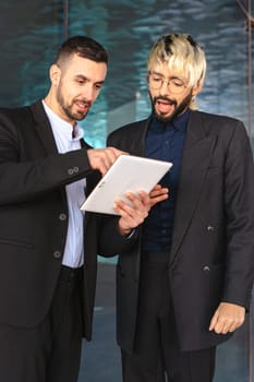 Two young businessmen in formal suits ,looking at a tablet and smiling while working together in a stylish modern office with reflective surfaces and a professional atmosphere. Vertical