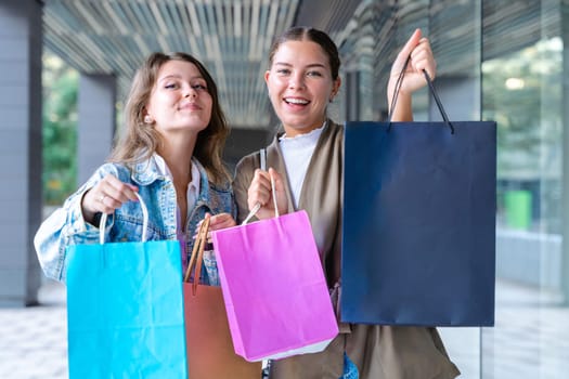 Two cheerful Caucasian women looking at the camera and smiling while holding colorful shopping bags. Capturing joyful and fun shopping moments in an urban outdoor setting.