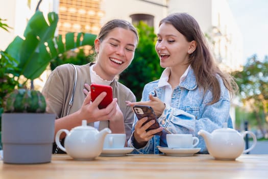 Two teenage girls share a fun moment at an outdoor cafe, smiling and pointing at smartphones. Surrounded by tea cups and sunshine, they enjoy friendship and technology together.