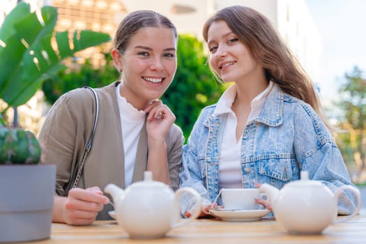 Portrait Two young Caucasian women smiling and looking at the camera while enjoying tea outdoors. Relaxed and cheerful moment of friendship, leisure, and conversation on a sunny day in the city.