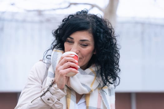 Caucasian woman enjoys a warm drink from a red cup while standing outdoors. She wears a soft scarf and light jacket, suggesting a cool day. Calm and cozy moment captured in natural daylight.