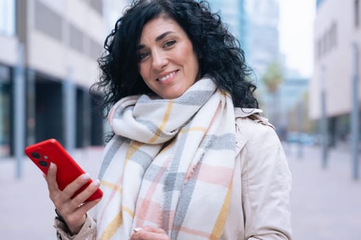 Happy caucasian woman standing outdoors in urban setting,wearing coat and scarf,using her red smartphone.She is smiling and relaxed,enjoying a casual moment on a cold day in the city looking at camera