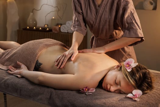Therapist giving relaxing back massage in cozy spa ambiance with petals, soft towel, and soothing light.