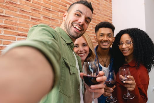 Multicultural group of young friends smiling while taking a selfie and toasting with glasses of red wine during a cozy indoor dinner, capturing joyful moments, connection, and celebration at home.