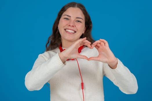 Happy young woman in white sweater and red headphones smiling while making a heart shape with her hands against a blue background, representing kindness, connection, love, and positive emotions.