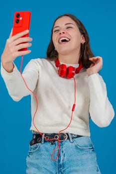Vertical image of a happy young woman taking a selfie with a red smartphone and headphones. She smiles confidently in front of a blue background, enjoying a casual and modern digital moment. Vertical