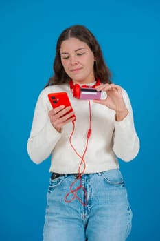 Happy young woman in a white sweater and red headphones smiling while showing a credit card to the camera and holding a red smartphone, against a blue background, symbolizing online shopping. Vertical